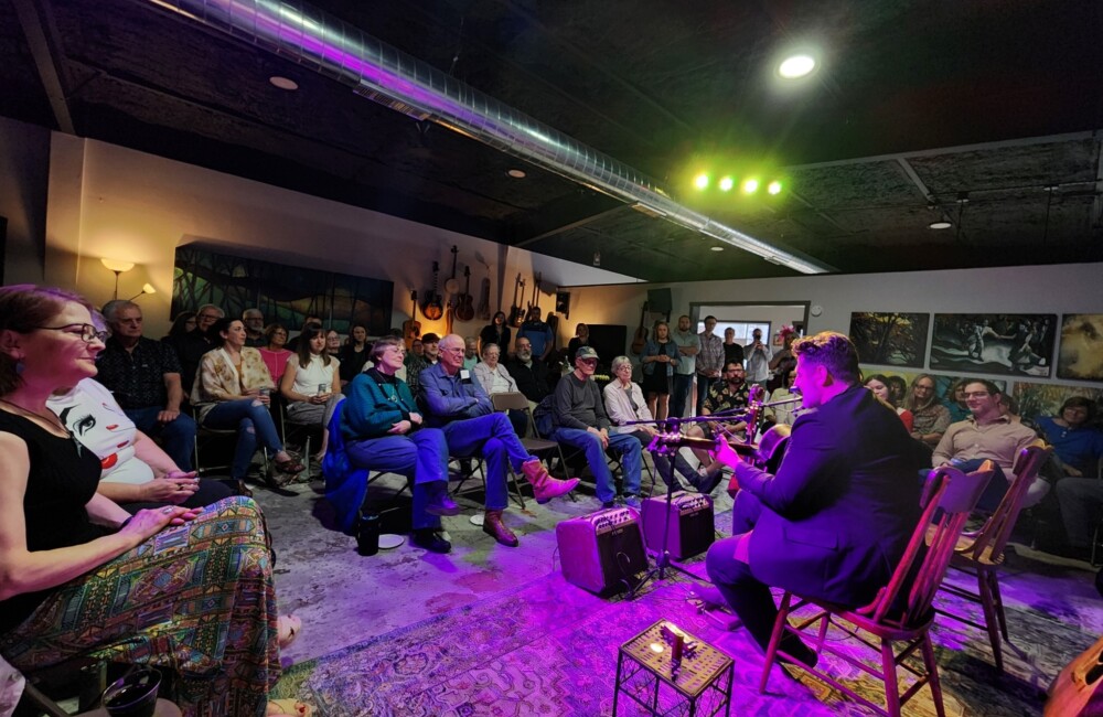 Bathed in purple light, a guitarist performs for a crowd gathered in a gallery space.