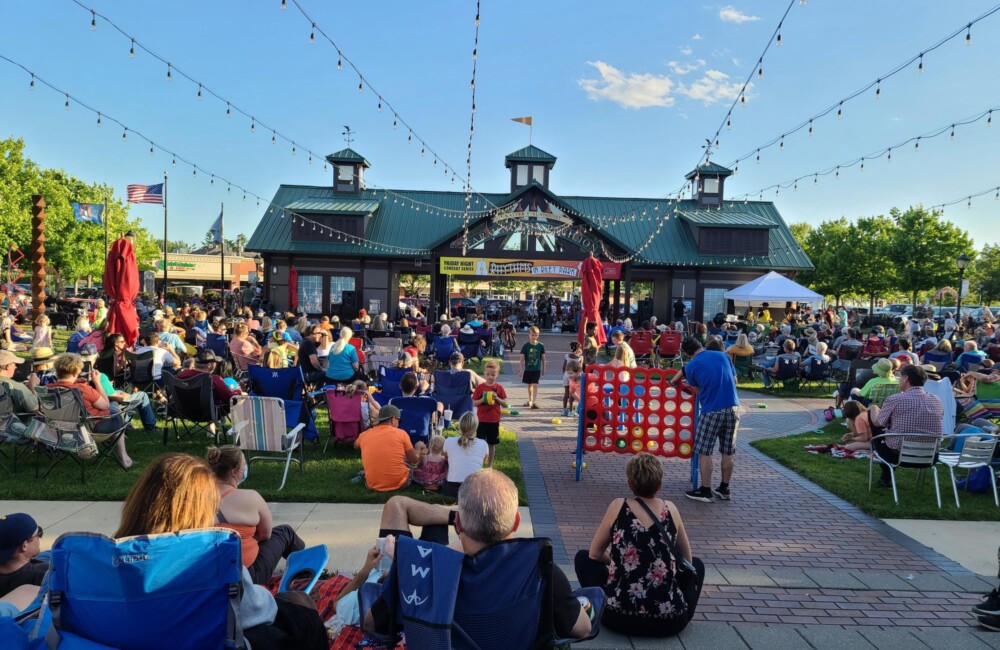 People sit in chairs and play lawn games in a large square anchored by a pavilion structure.