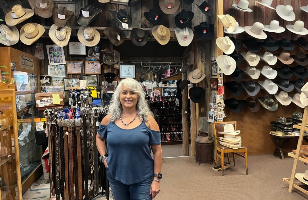 A woman stands proudly beside fixtures displaying Western-style hats, belts, and memorabilia.