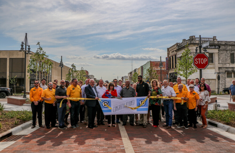 A large group of people stand in the center of a newly paved street to cut a ribbon commemorating the completion of a streetscape project.