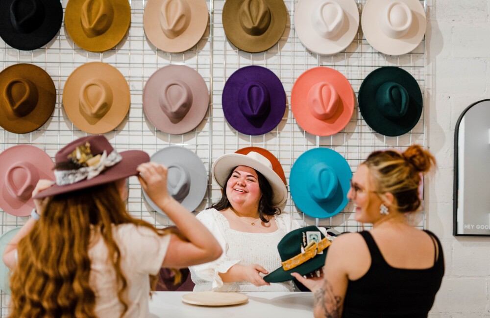 Women shop for hats in a brightly lit shop featuring a wall of colorful brimmed hats.