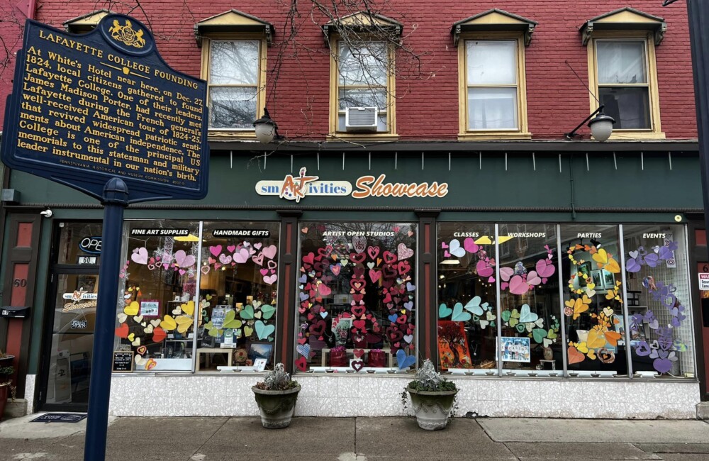 A mosaic of colorful paper hearts fills the ground floor windows of an art supply store; in the foreground, a historic marker for Lafayette College is visible.