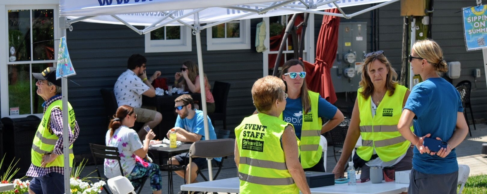 People wearing fluorescent vests stand around a table under a tent with the words "downtownsykesville.com"