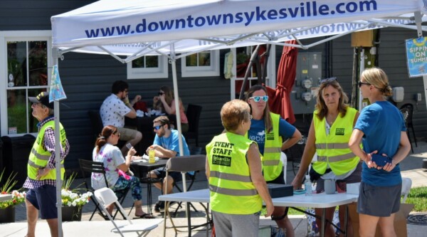 People wearing fluorescent vests stand around a table under a tent with the words "downtownsykesville.com"