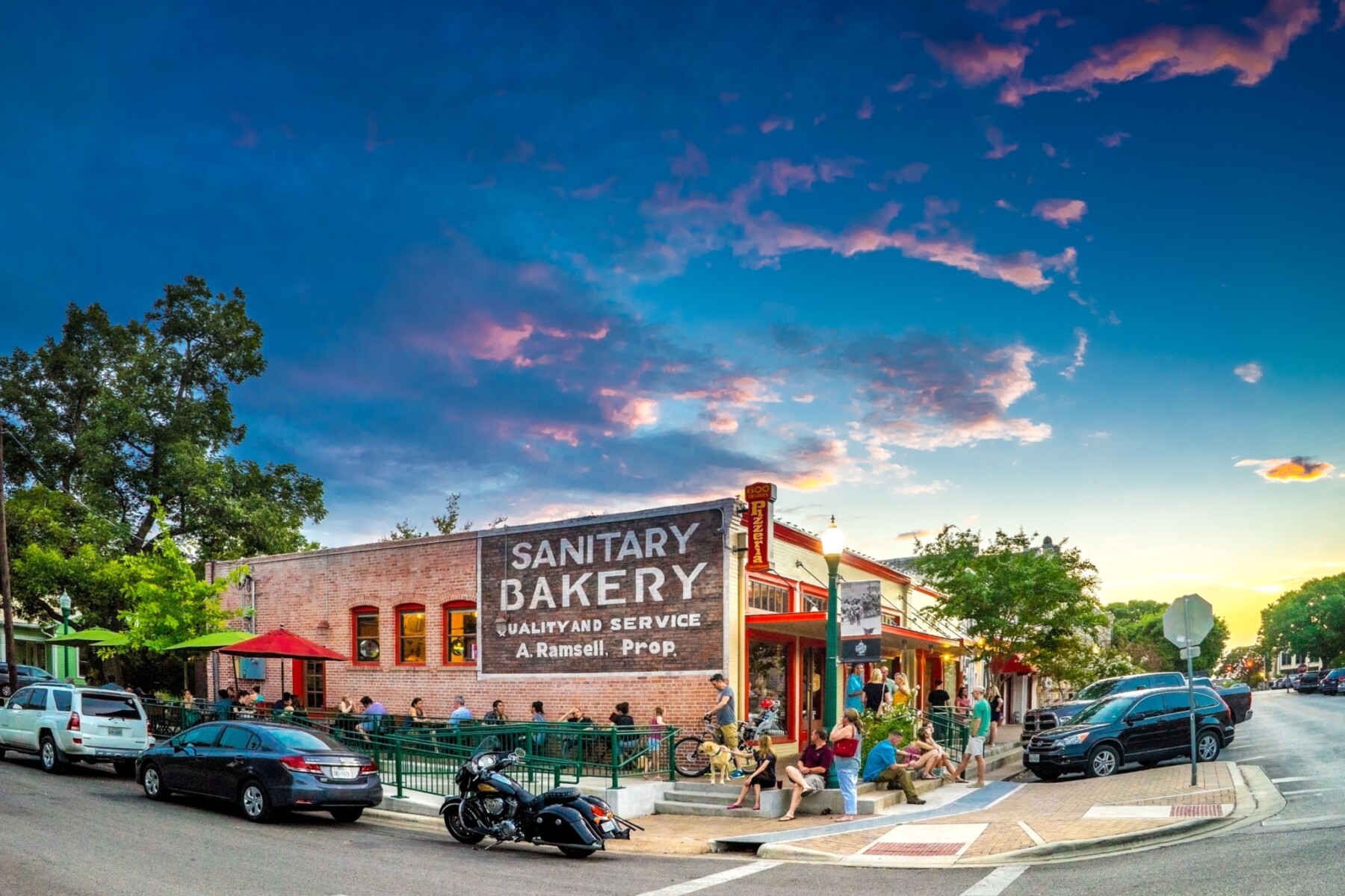 Historic brick building on city corner where people gather outside in seating area. The side of the building has an old-fashioned painted sign reading, "Sanitary Bakery Quality and Service A. Ramsell Prop."