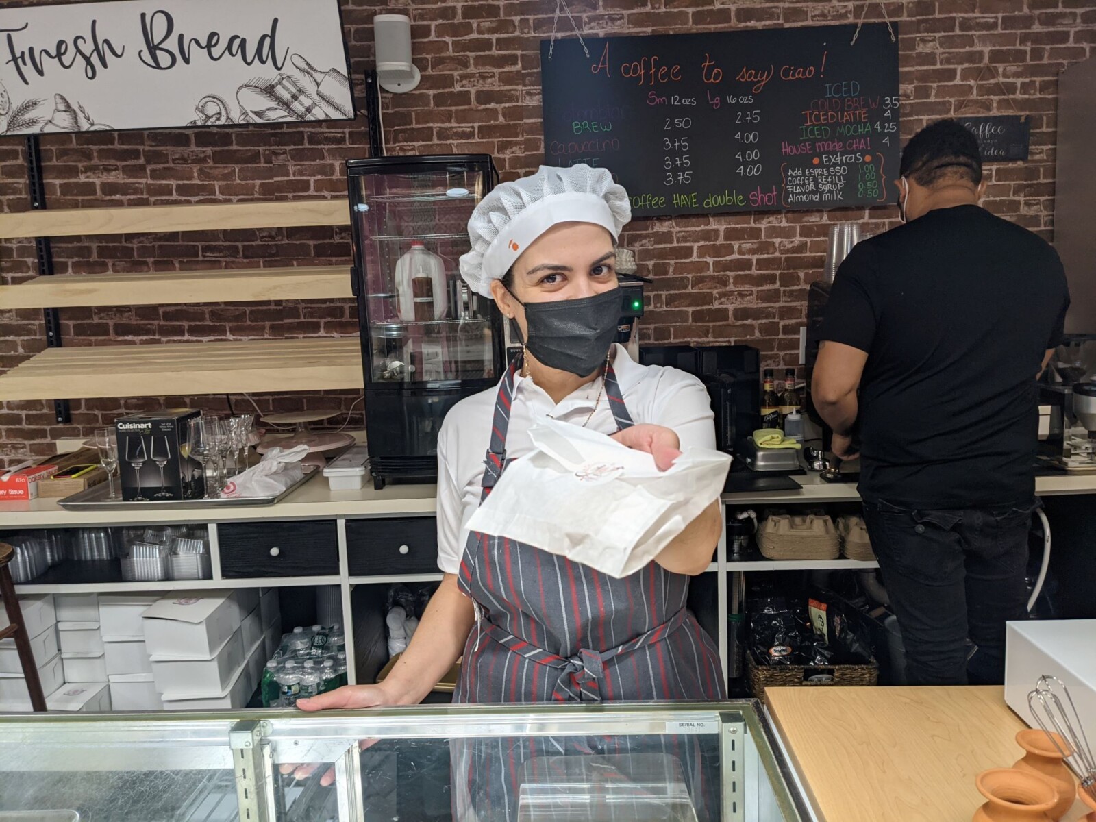 Woman behind bakery counter wearing chef's hat, apron, and face mask holds a pastry bag toward the camera