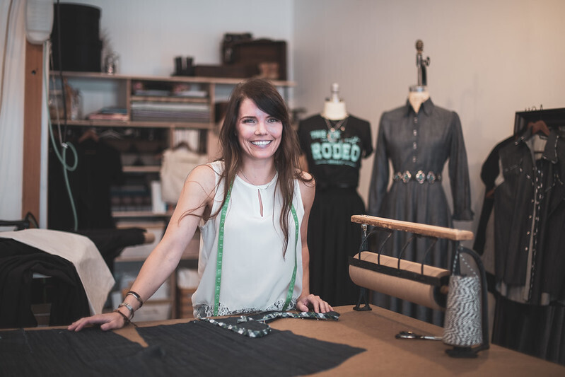 Woman with brown hair wearing white tank top smiles at camera while standing at desk where she is measuring fabric. In background, clothing hangs on mannequins.