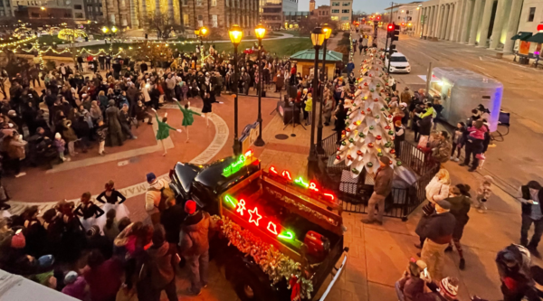 Crowd of people gather in outdoor pedestrian area watching dancers perform. A white Christmas tree and truck decorated with holiday lights sit nearby.