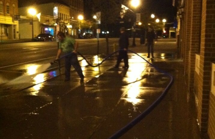 Two men holding a firehouse wash a downtown sidewalk