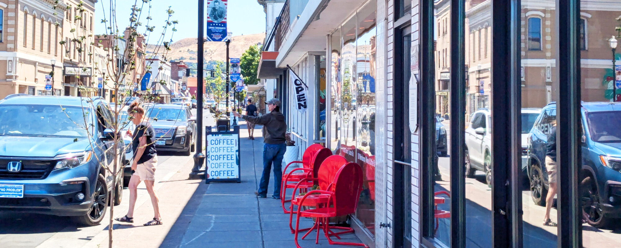 People walk along a sidewalk and a woman standing near a sign promoting a coffee business waves; bright red chairs offer seating along the exterior facade of the building.