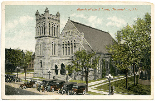 A colorized postcard depicting a church made of stone with model-T cars parked in front.