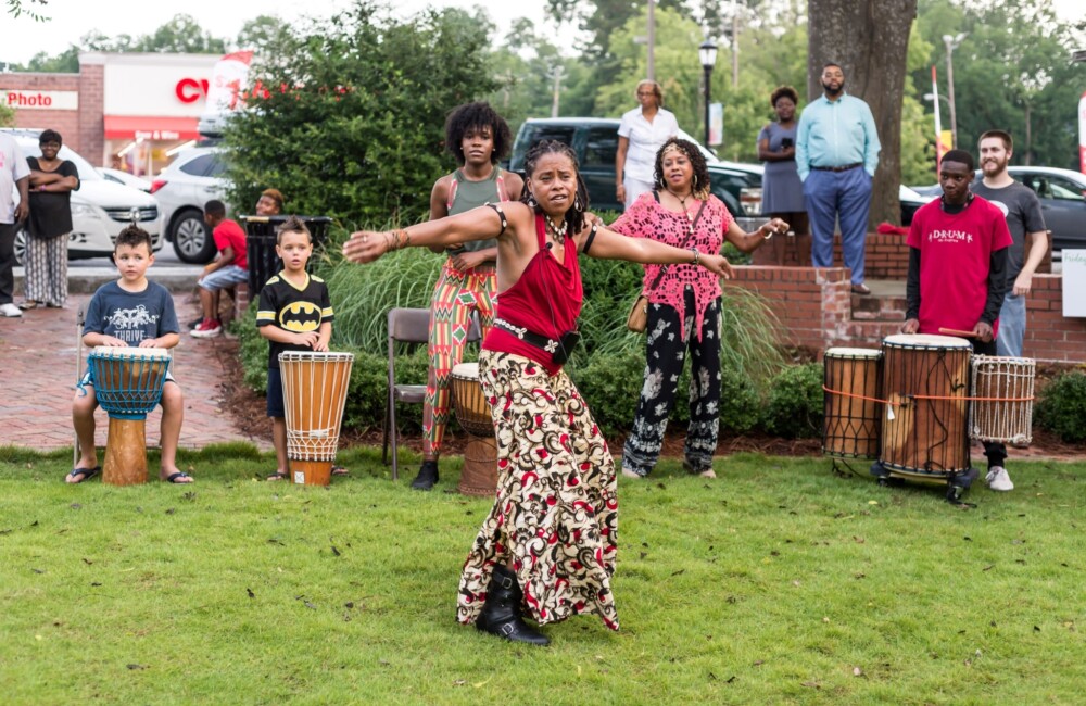 A young woman dances in the center of a drum circle while behind her young people play drums.