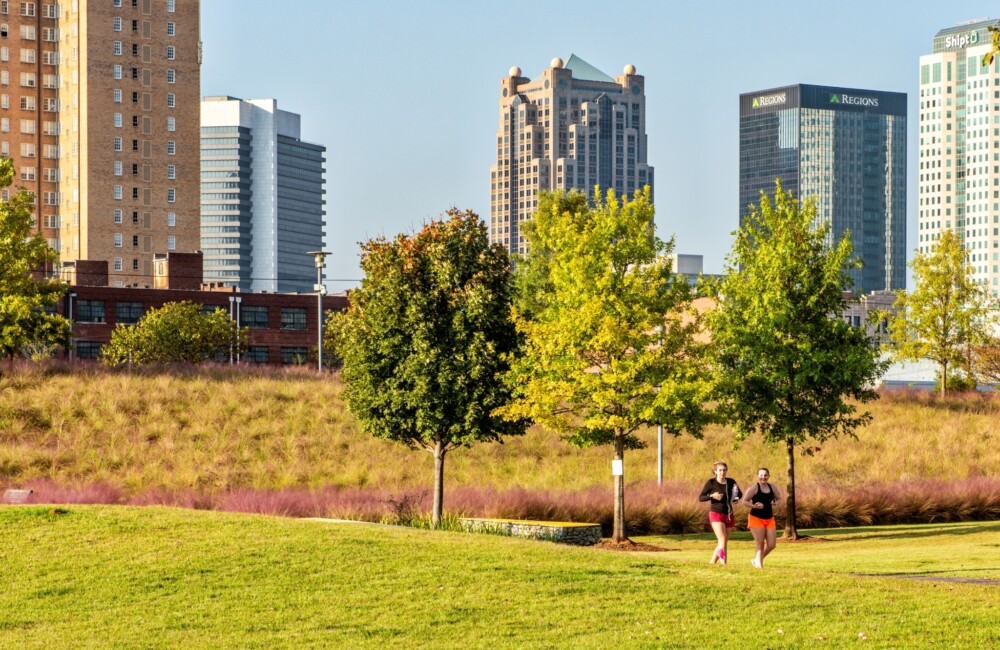 Two women jog through a lush park while in background the downtown Birmingham skyline is visible.