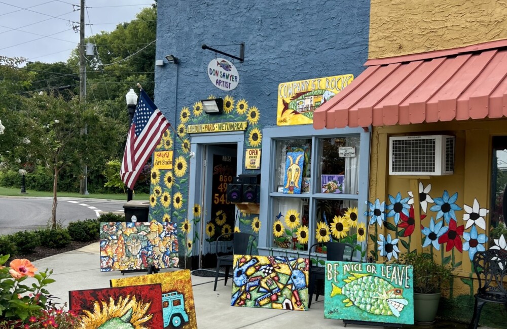 Colorful paintings are displayed on easels set up on the sidewalk outside of an art gallery.