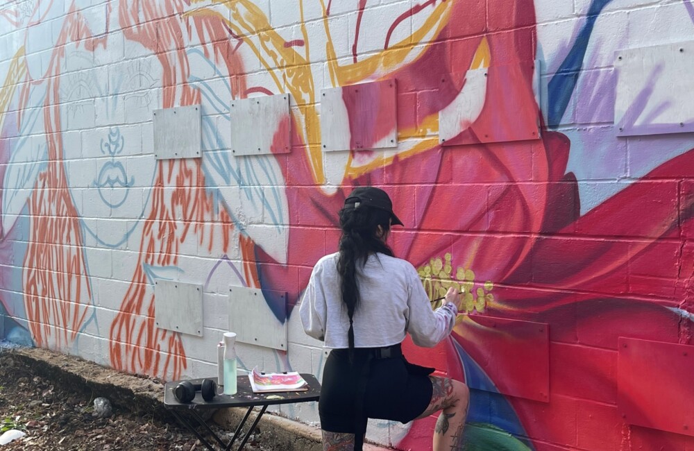 A woman kneels to apply paint to a mural in progress.