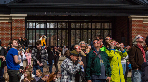 People wearing eclipse glasses stare up at the sky in front of a large brick and glass building