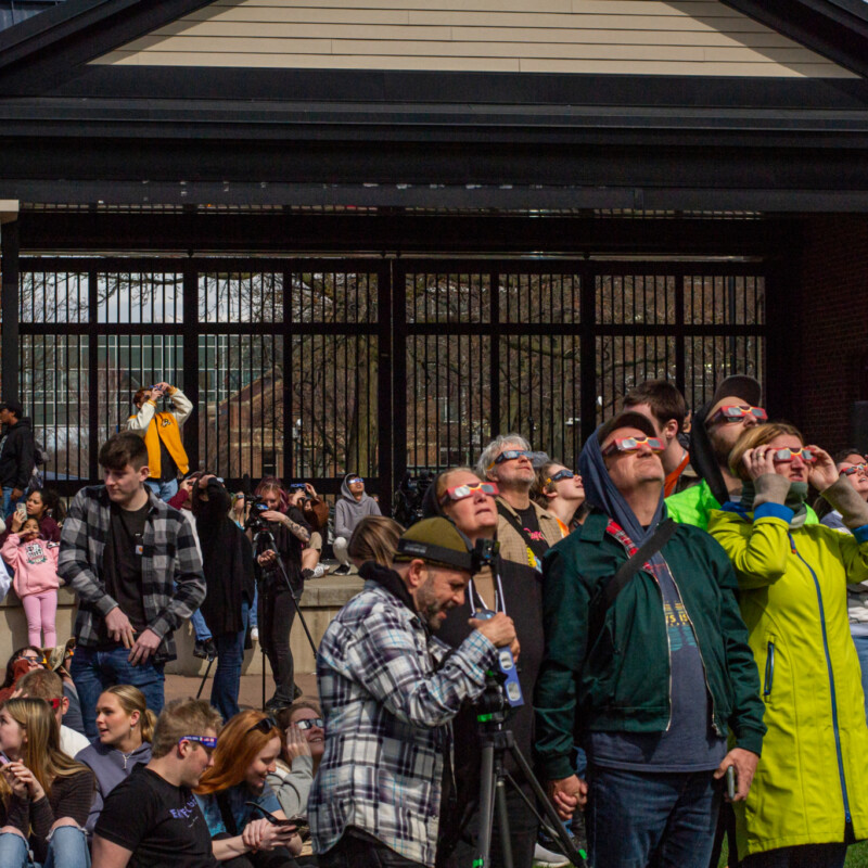 People wearing eclipse glasses stare up at the sky in front of a large brick and glass building