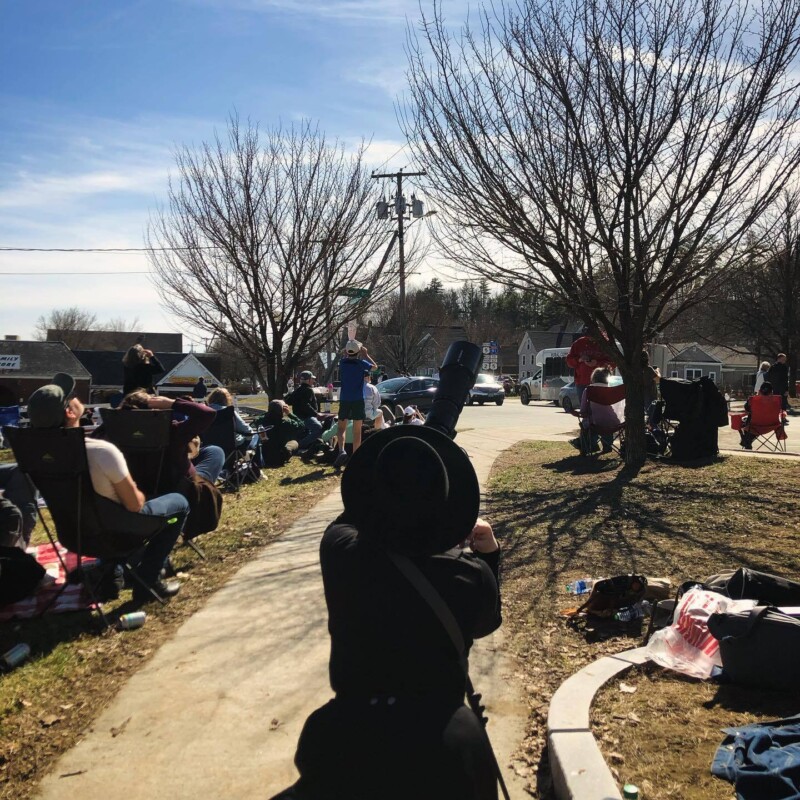 A person looks up at the sky through a telescope while others look through eclipse glasses. Bare trees and a sidewalk in the background.