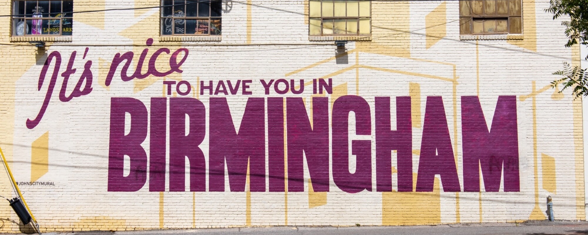 Mural on the exterior wall of a building reads "It's Nice to Have you in Birmingham" in a deep magenta color.