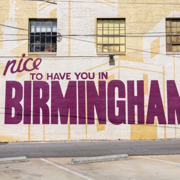 Mural on the exterior wall of a building reads "It's Nice to Have you in Birmingham" in a deep magenta color.