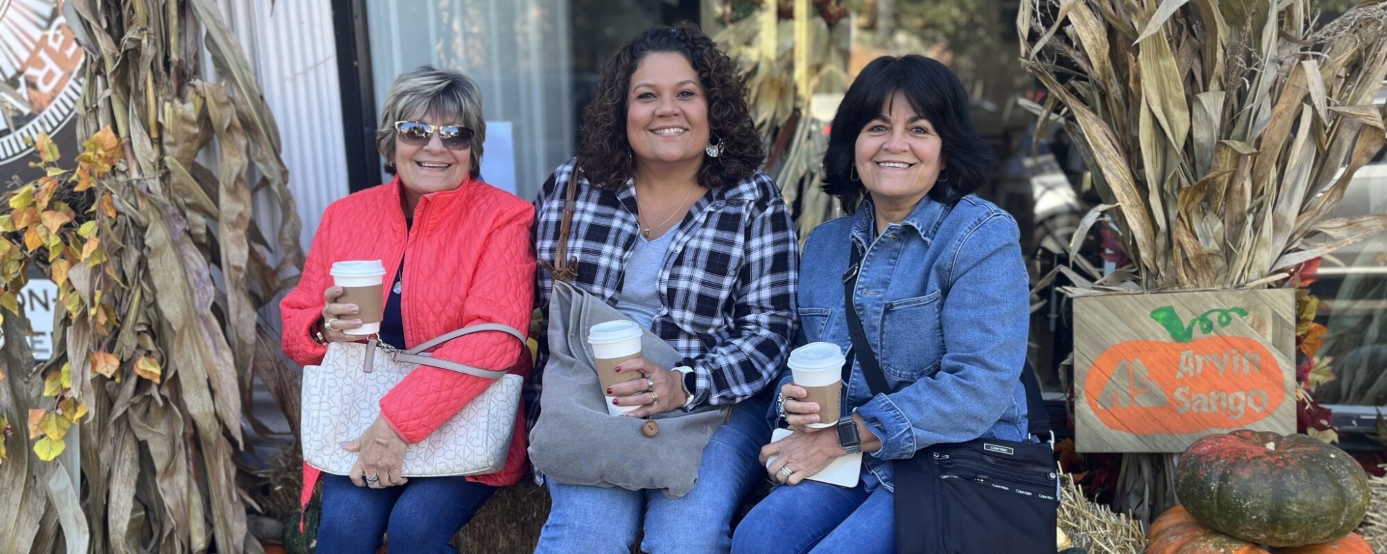 Three women hold cups of coffee while sitting on a hay bale.