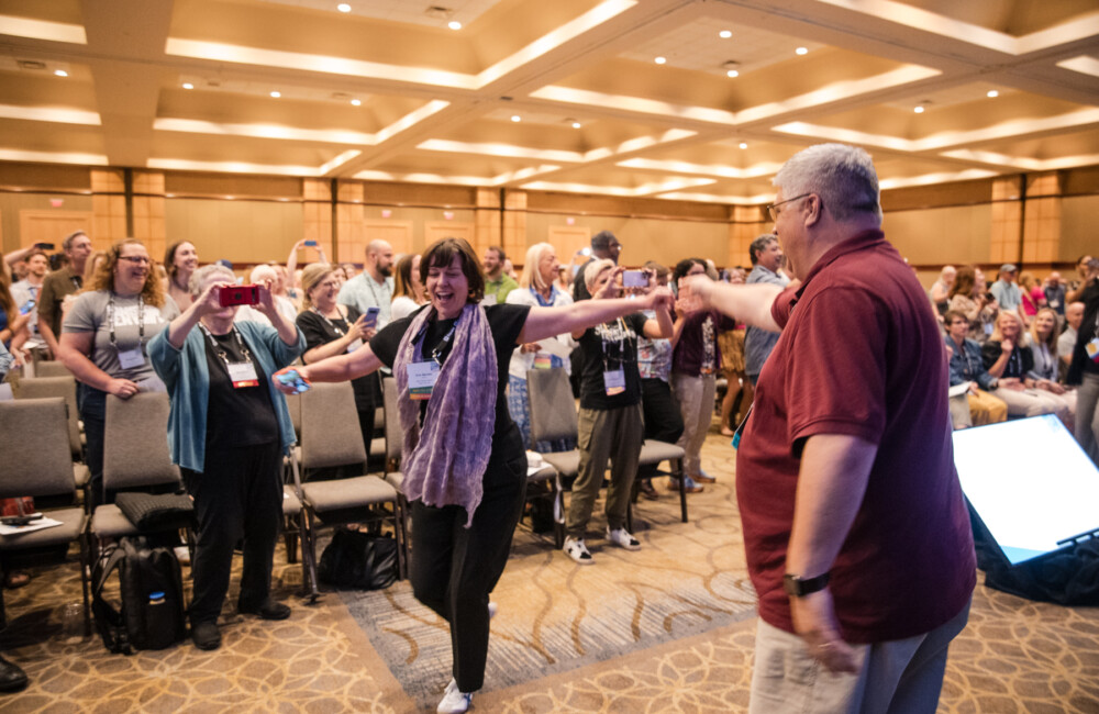 Two people dance inside a large hotel ballroom; behind them people are standing and dancing.