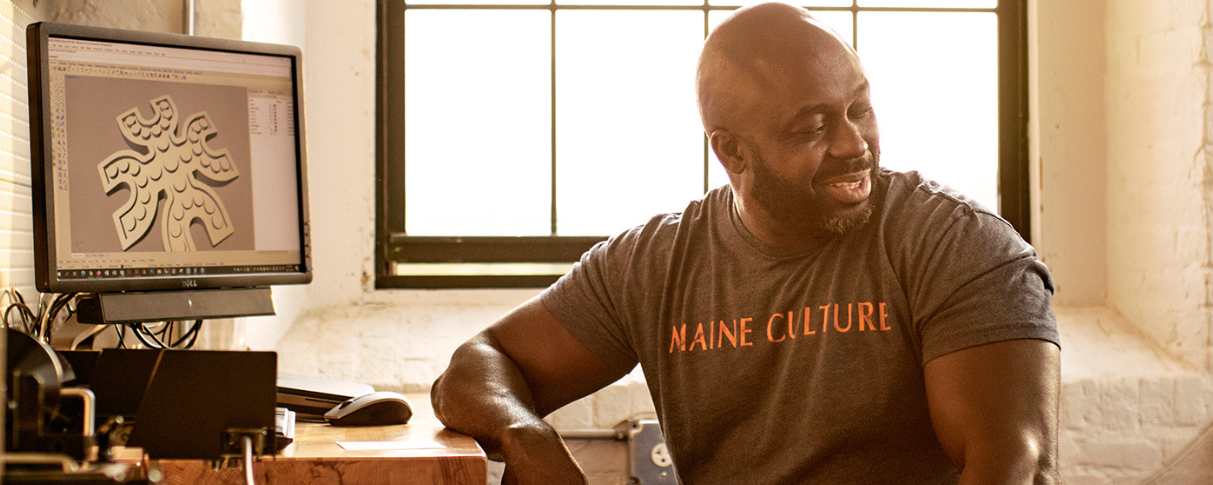 Man in t-shirt reading, "Maine Culture," sits at desk in front of computer.