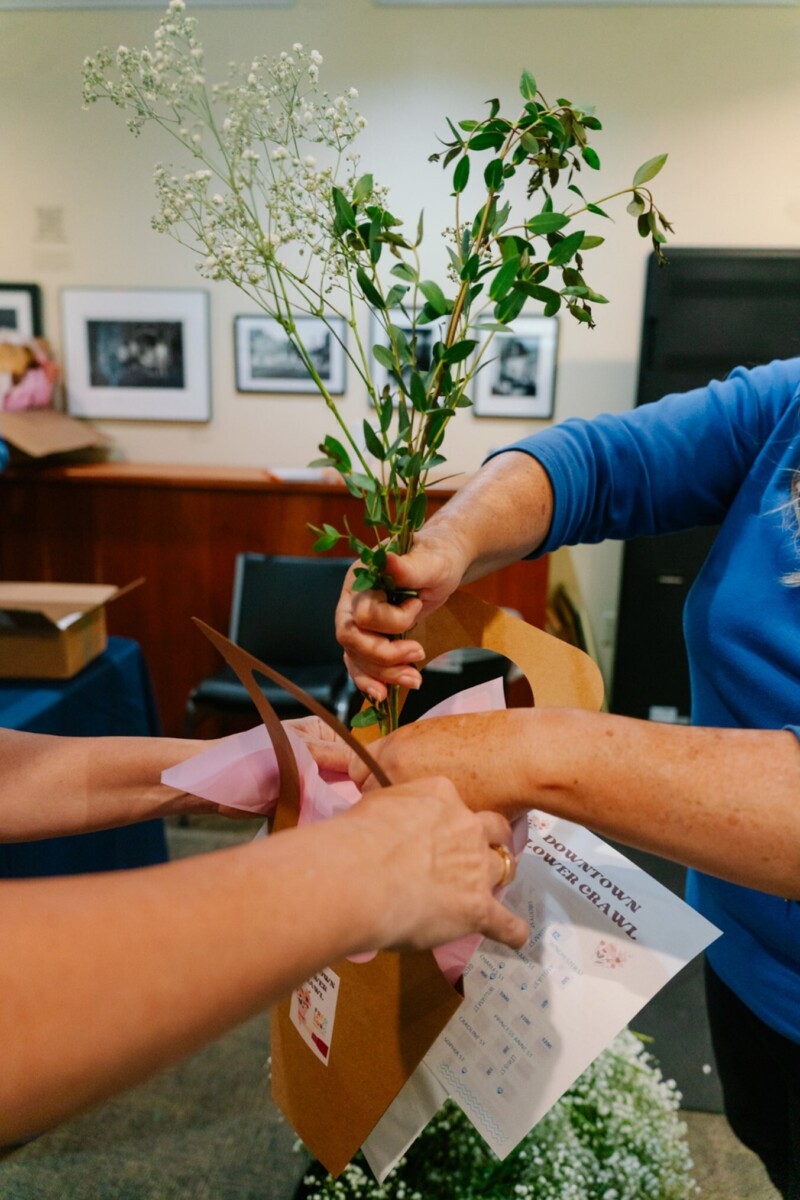 A person holding some stems of greenery helps another person set up their flower carrier. The other person is also holding the map of the crawl.