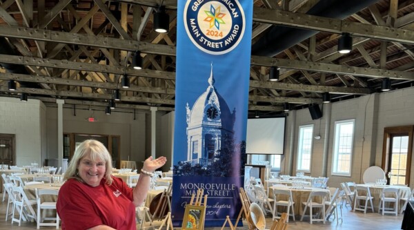 Una mujer señala con entusiasmo una pancarta vertical que celebra que Monroeville Main Street ha sido galardonada con el premio Great American Main Street 2024.