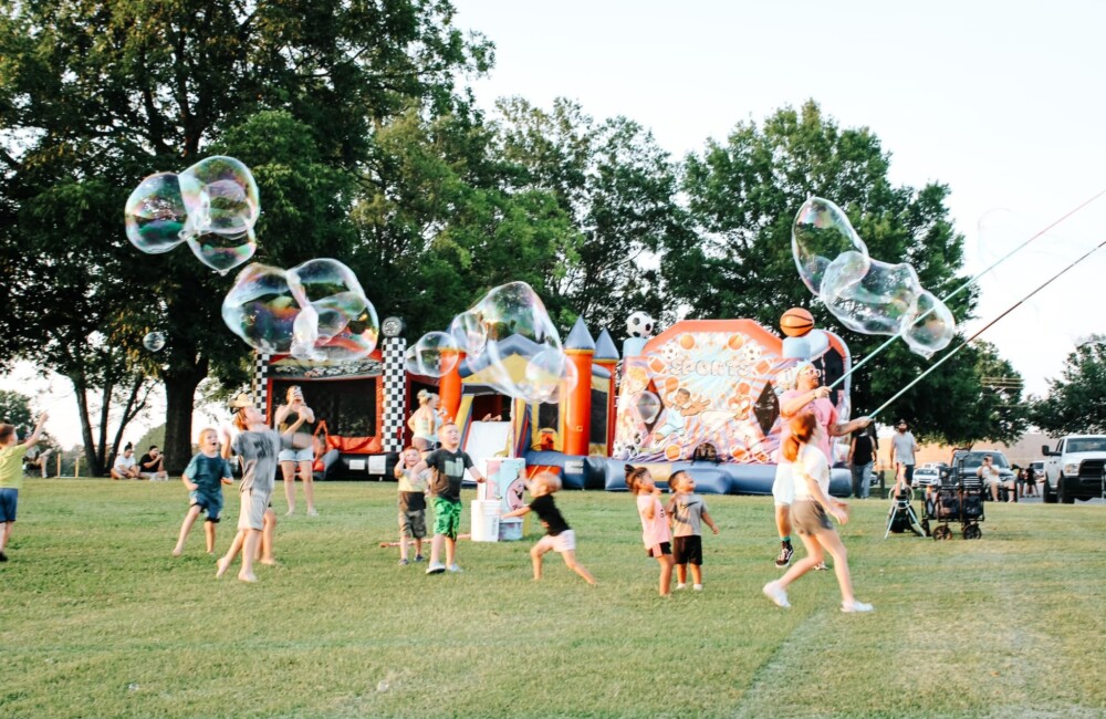 Children play with bubbles on grassy field in front of bouncy house.