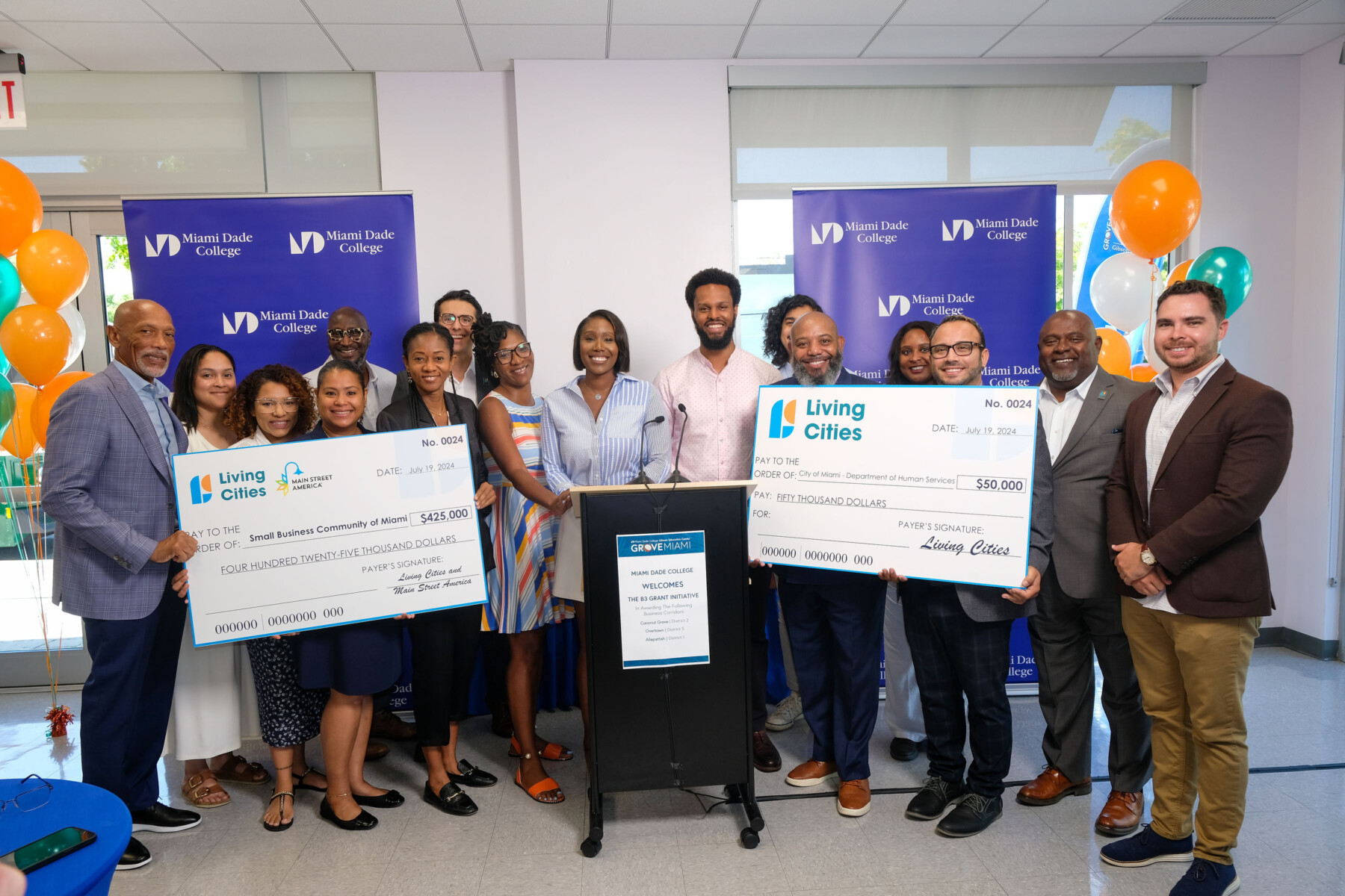 A group of people pose for photos in front of conference room while holding two large checks.