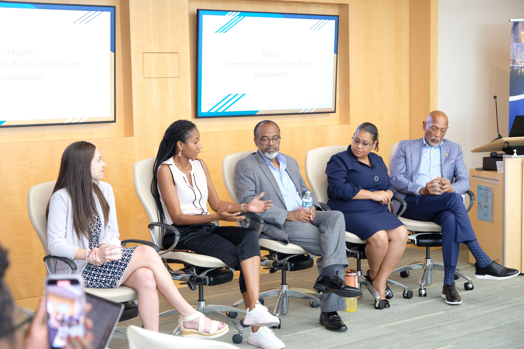 A panel of five speakers sit in chairs in front of conference room.