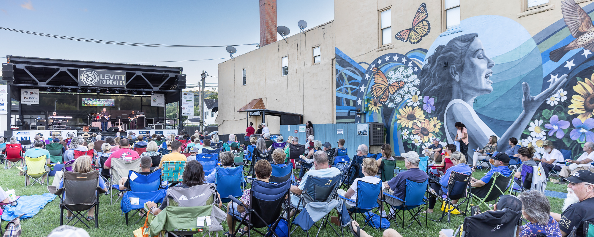 Group of people sit on lawn chairs on grassy lawn while listening to live music on outdoor stage next to mural on exterior of historic building.