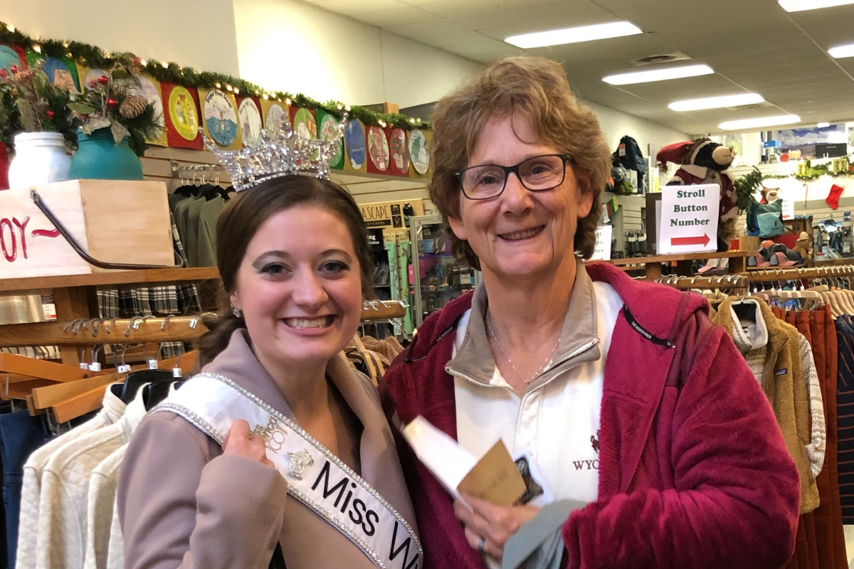 Two women smile in front of clothing racks in small business. The woman on the left wears a tiara and a white sash reading, "Miss Wyoming." The woman on the right is wearing a red sweatshirt over a white shirt.