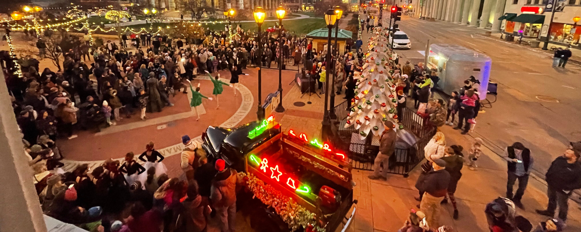 Crowd of people gather in outdoor pedestrian area watching dancers perform. A white Christmas tree and truck decorated with holiday lights sit nearby.