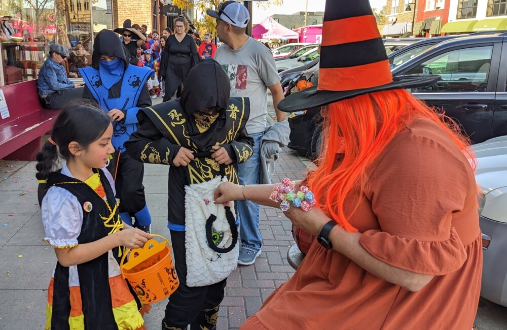 Kids dressed up in Halloween costume open their bags for trick or treat candy from a woman dressed as an orange witch