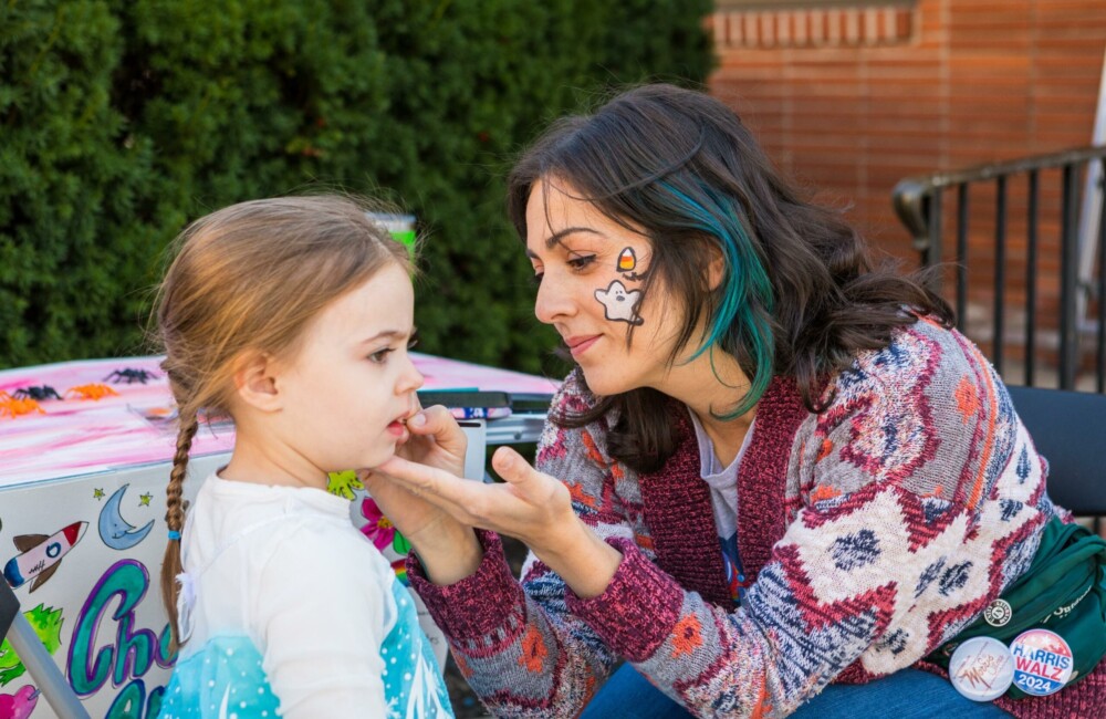 A little girl wearing an Elsa costume has her face painted by a woman