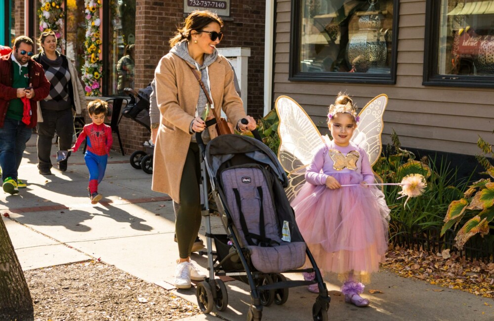 A little girl wearing a pink fairy costume walks beside her mother pushing an empty stroller. Behind them is a little boy wearing a spider man costume.