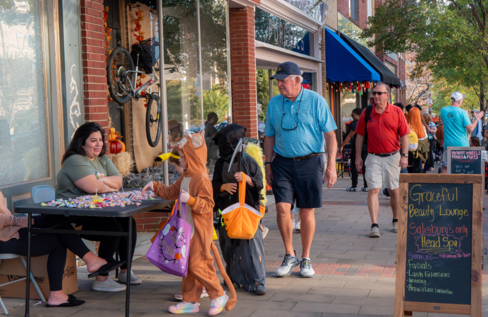 People walking down a downtown sidewalk in Halloween costumes, a table outside a small business is handing out candy