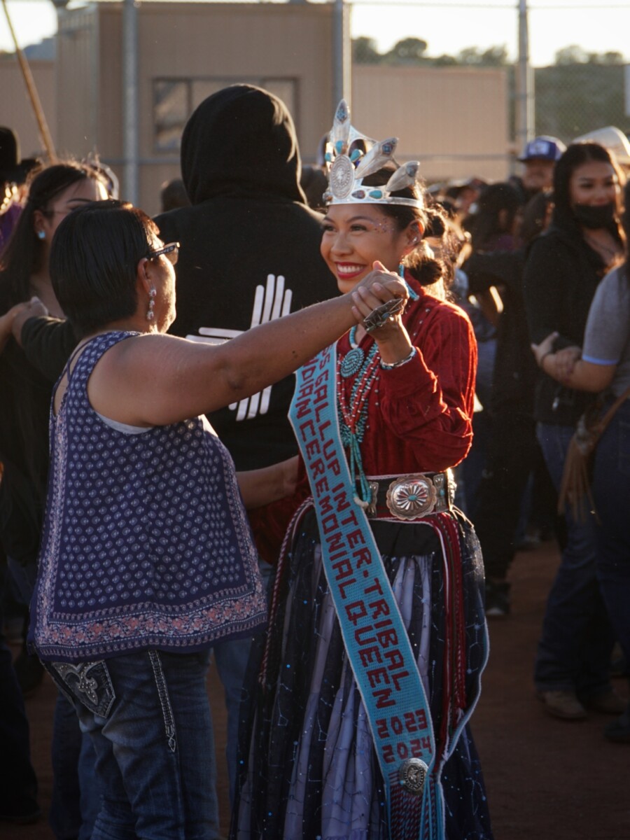 Two women smile and dance in outdoor music venue. One of the women wears traditional Native American dress.