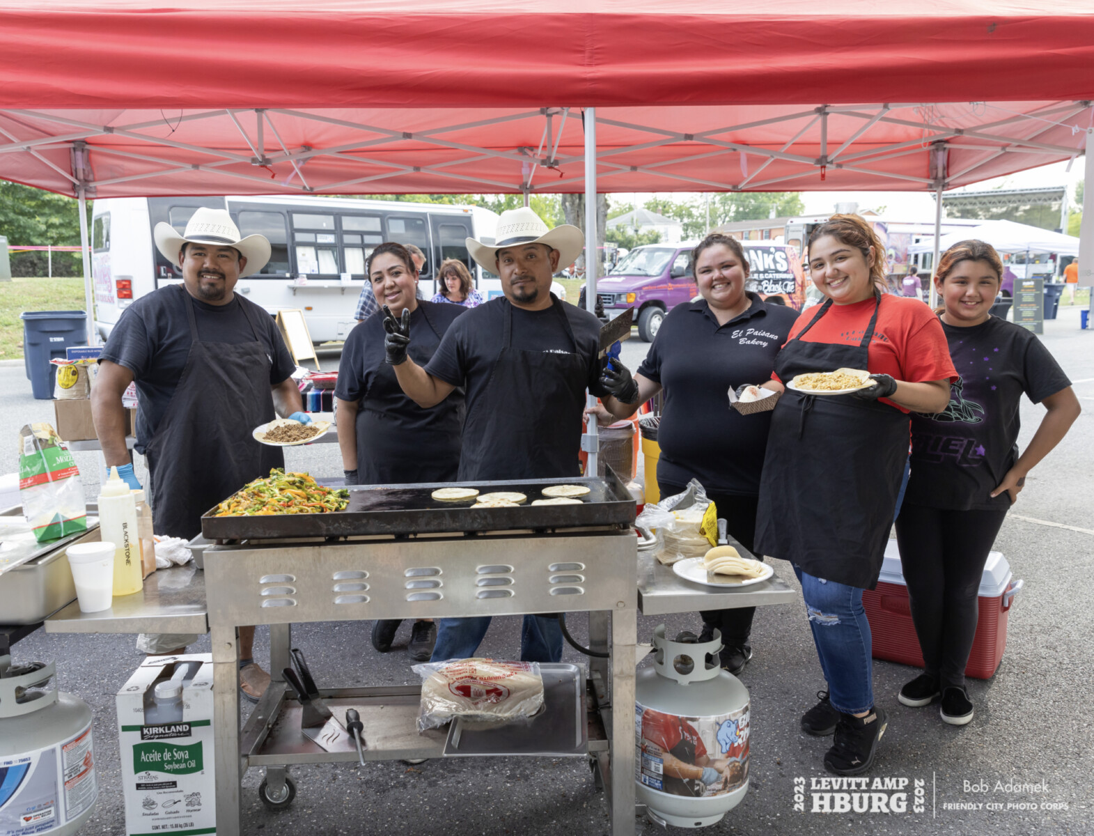 Group of six people wearing black aprons smile and hold plates of food in front of grill under outdoor food vendor tent.