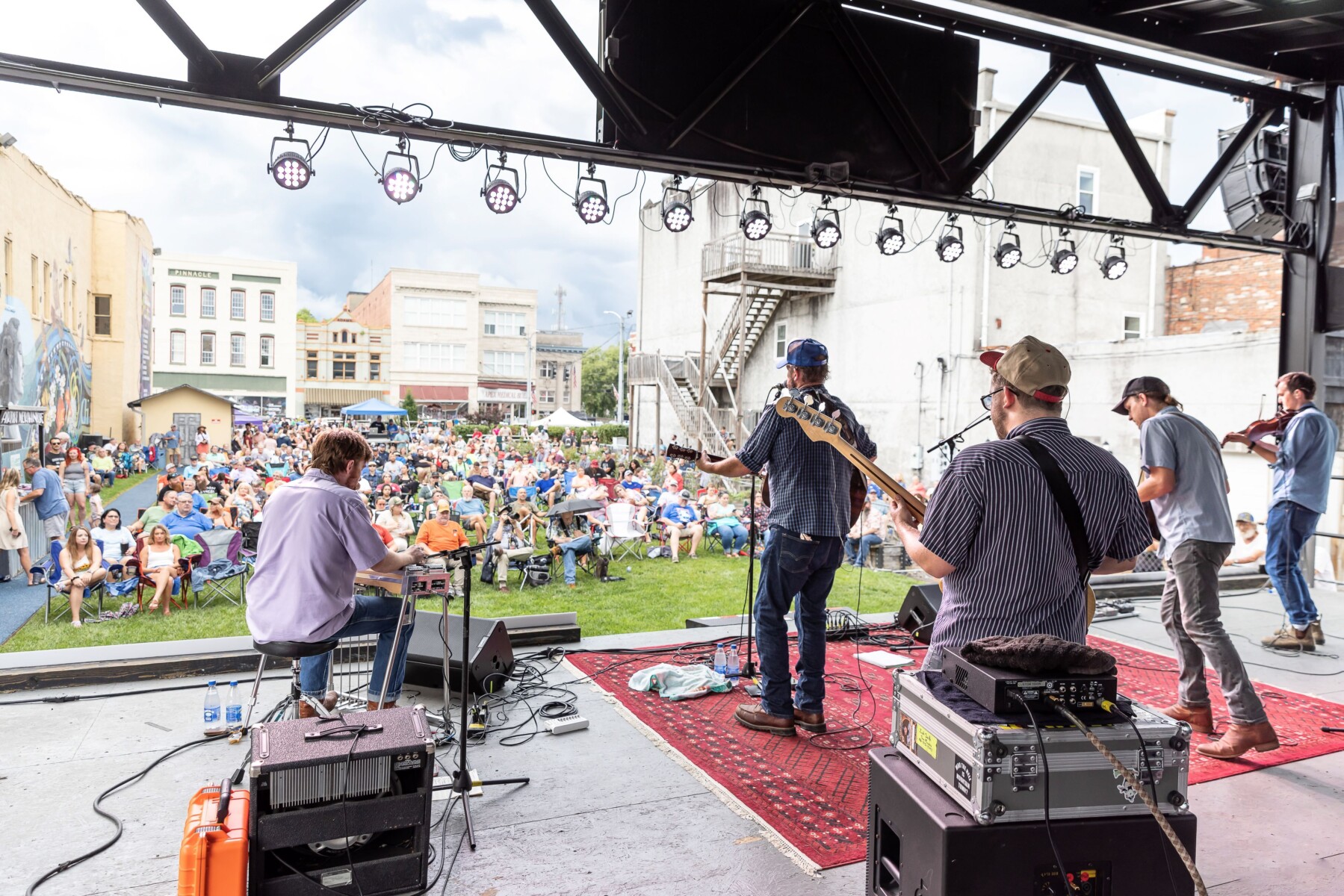 Band plays on outdoor stage in front of large crowd sitting on lawn.