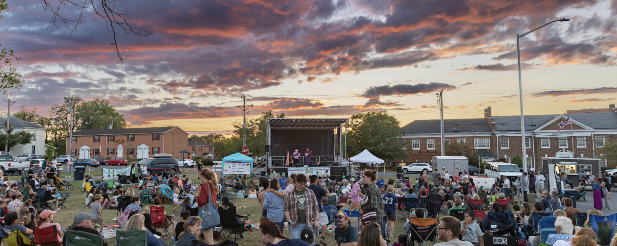 At sunset, group of people gather on lawn in front of stage.