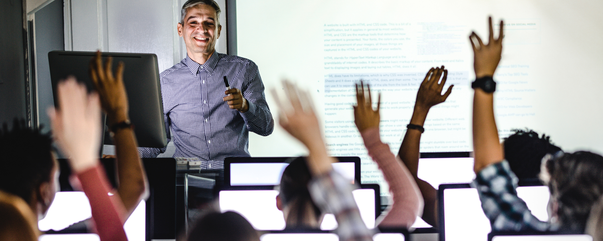 Students in a computer lab raise their hands while a smiling adult professor faces them.