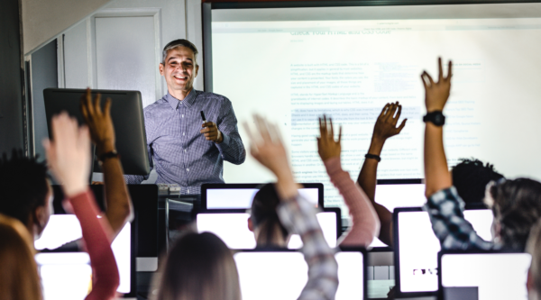 Students in a computer lab raise their hands while a smiling adult professor faces them.