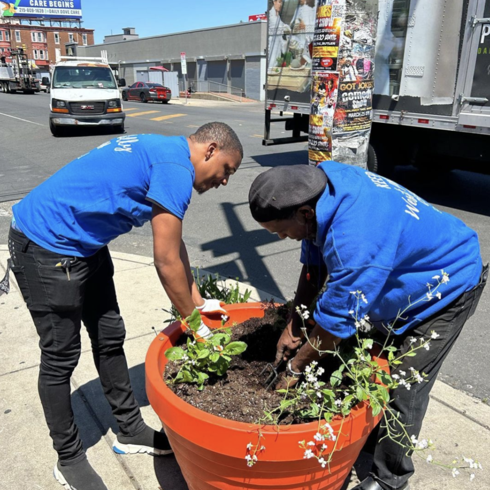 Two men wearing bright blue shirts place plants into a vermillion-colored planter.