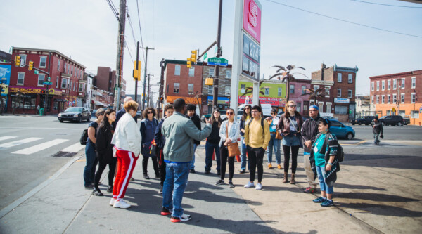 A group of people stand on a busy city street corner, listening to a tour guide speak about the neighborhood.