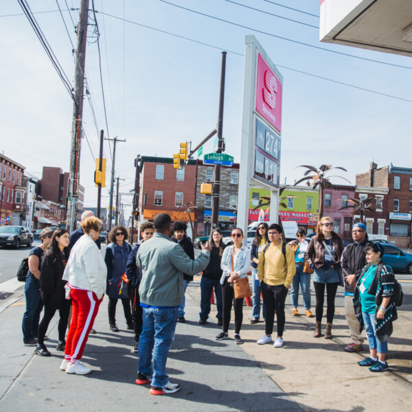 A group of people stand on a busy city street corner, listening to a tour guide speak about the neighborhood.