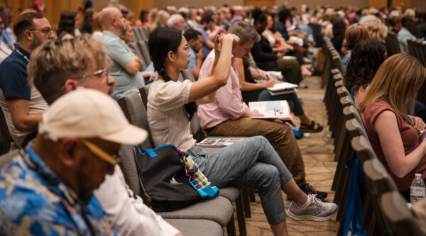 People attending a large session take notes while listening to a presenter (out of frame).