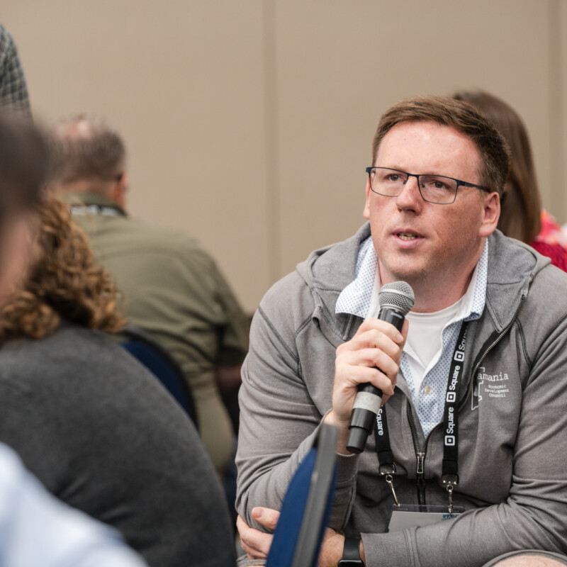 A man sitting in a classroom-style audience speaks into a microphone.